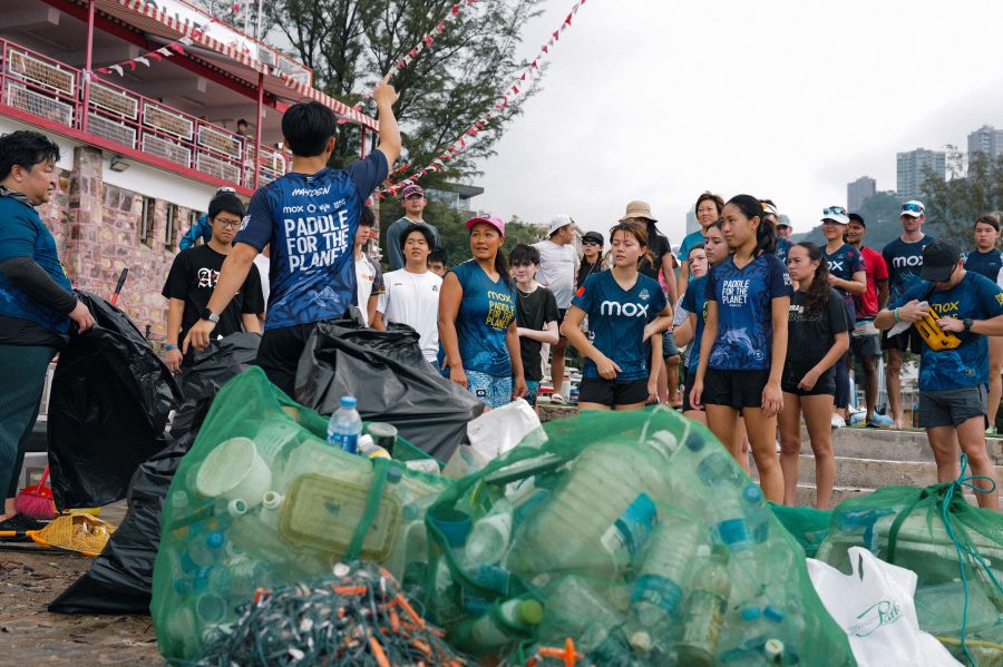 Group of volunteers at a cleanup event, standing around large piles of plastic bottles and bags of trash on a waterfront area.