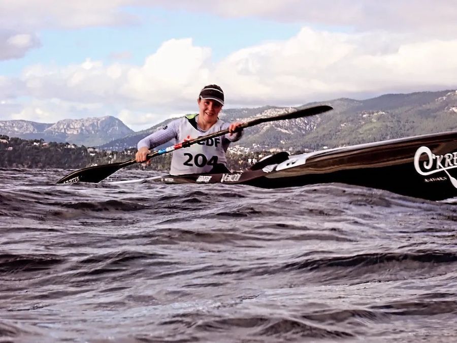 Kayaker wearing bib 204 paddles a black racing kayak through choppy water with a coastline and hills in the background.