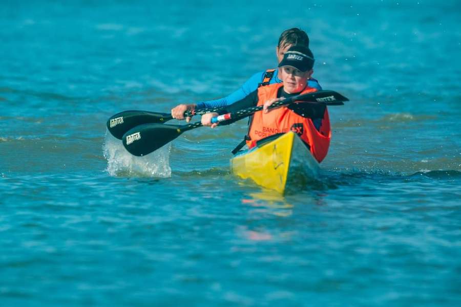 Person wearing an orange life jacket paddling a yellow kayak on blue water with a double-bladed paddle.