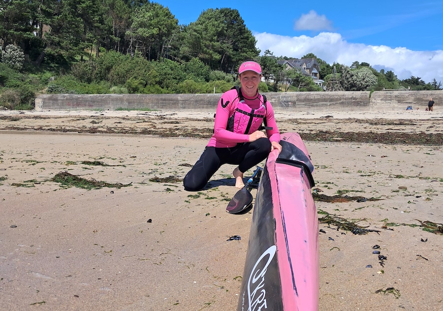 Person in a pink wetsuit kneeling on a sandy beach beside a pink surfboard, with green trees and blue sky in the background.