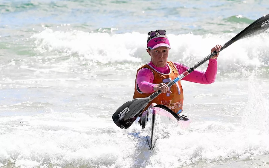 Female kayaker paddling through breaking waves, wearing a pink long-sleeve top, orange vest, and cap with sunglasses perched on it.