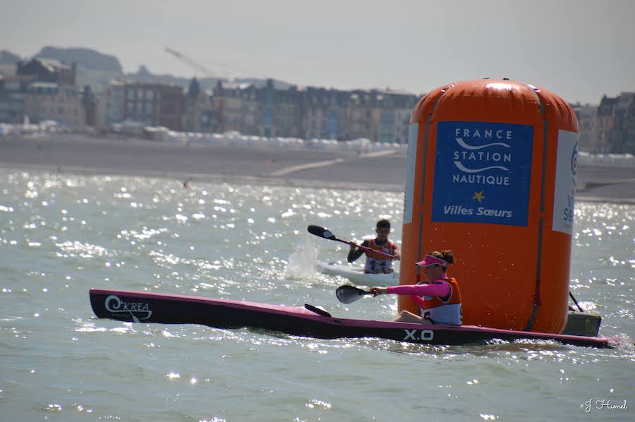Two kayakers race in a pink and black kayak beside a large orange buoy on glistening coastal water, with a town in the background.