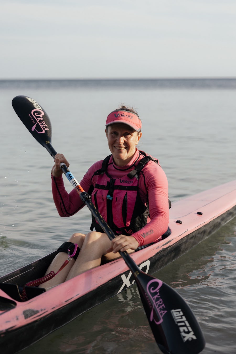 Woman in pink kayaking on calm water, smiling as she paddles a pink kayak with a black double-bladed paddle.