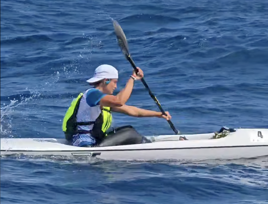 Kayaker wearing a white hat and neon life jacket paddling a white kayak on the blue ocean, mid-stroke with the paddle raised above water.