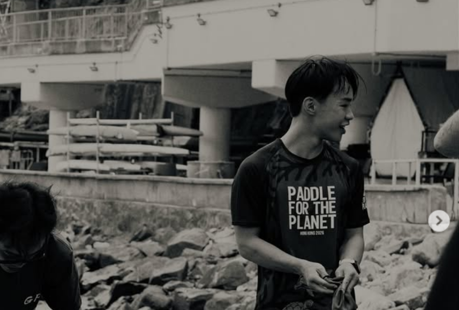 Young man in a 'Paddle for the Planet' shirt stands on a rocky shoreline near a marina, smiling. (informative)