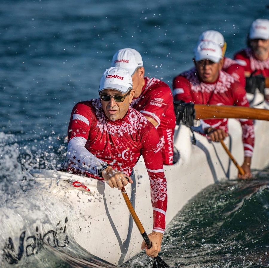 A team of rowers in red uniforms paddling a white boat through choppy water, wearing white caps labeled Singapore.
